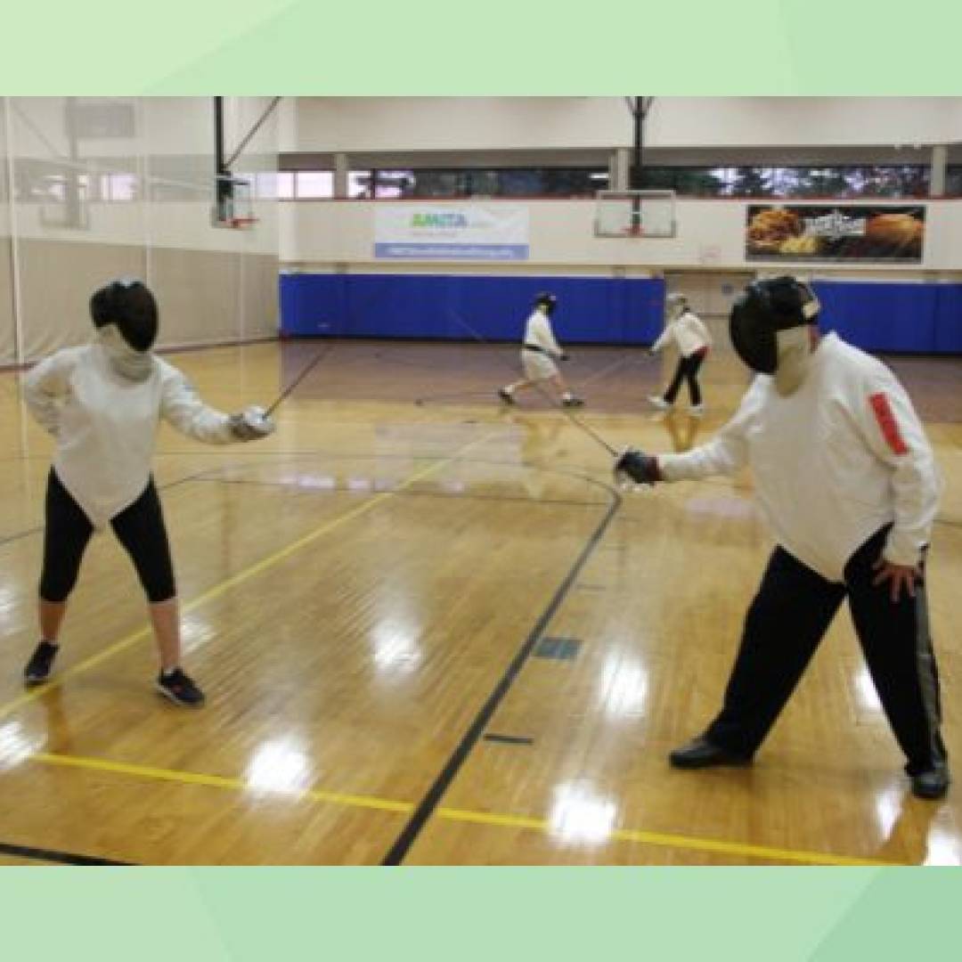 Adults participating in fencing practice, wearing protective gear and sparring with foils in a gym setting.